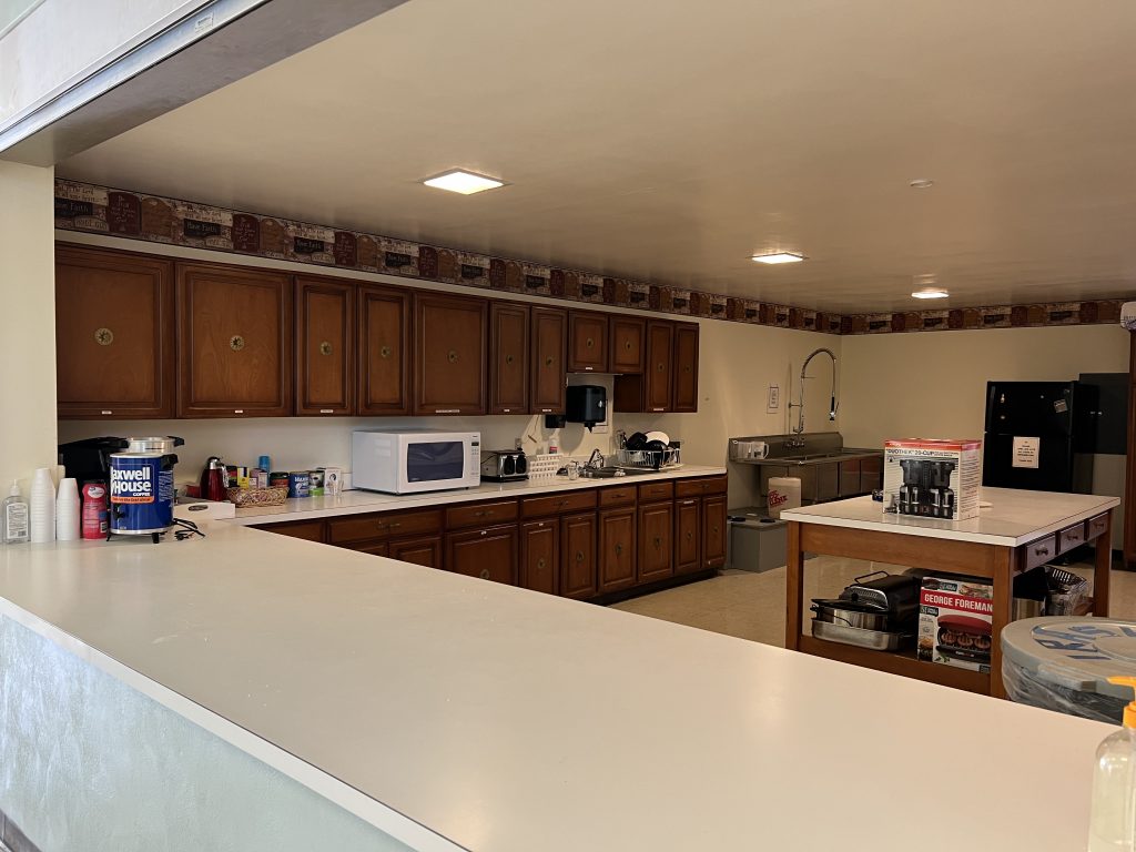 Image depicts kitchen located in the Fellowship Hall, it includes counterspace, serving window, sink, and appliances. 